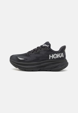 Black athletic shoes with mesh upper, thick cushioned sole, and prominent logo. Features laces and textured patterns for traction.