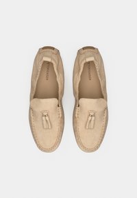 Mocassins en daim beige avec une semelle plate. Ils présentent un gland noué et des détails cousus autour des bords. Texture lisse, design minimaliste.