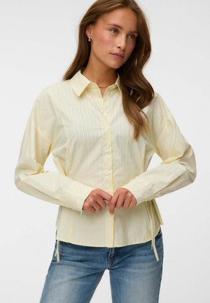 Femme aux longs cheveux bruns portant une chemise à rayures verticales jaunes et blanches boutonnée et un jean bleu, les mains jointes devant.