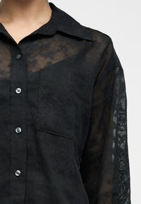 Κοντινό πλάνο ενός ατόμου που φοράει μαύρο διάφανο πουκάμισο με ανθικό μοτίβο, κουμπωτό, με αριστερή τσέπη στο στήθος και γιακά.