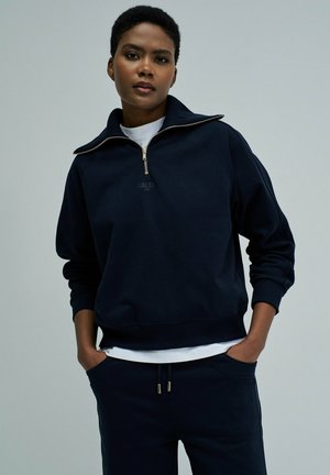 Jeune personne portant un sweat-shirt zippé bleu marine avec les mains dans les poches, debout devant un fond gris uni.