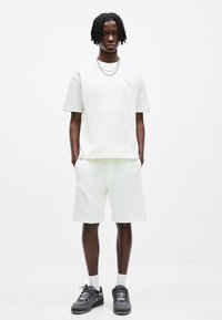 Weißes Baumwoll-T-Shirt und Shorts-Set mit lockerer Passform. Texturiertes Material, Rundhalsausschnitt und gesticktes Logodesign. Schwarze Turnschuhe mit grauen Akzenten.