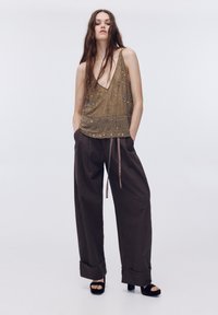 Gyöngyös olívazöld ujjatlan felső mély V-nyakú, laza barna, magas derekú nadrággal és fekete platform szandállal párosítva.