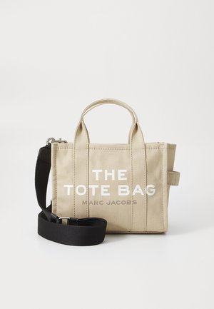 Beige auduma soma ar diviem augšējiem rokturiem un noņemamu melnu siksnu. Uz soma ir izcila balta teksts: "THE TOTE BAG MARC JACOBS."