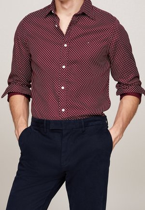 Mann trägt ein dunkelrotes, gemustertes Hemd mit hochgekrempelten Ärmeln und dunkelblaue Hose, die Hände in den Taschen.