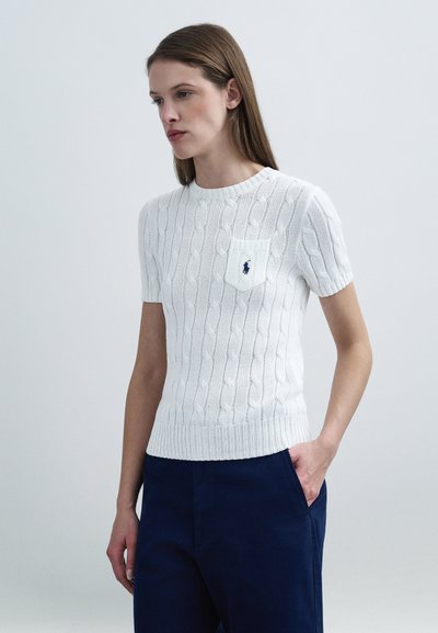 Pull blanc en maille torsadée avec un col rond, des manches courtes et une poche plaquée avec un petit logo bleu marine. Porté avec un pantalon sombre.
