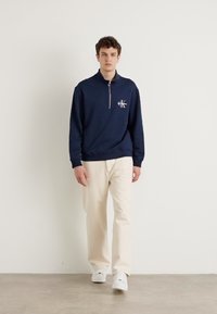 Felpa zip-up blu navy con colletto alto e logo ricamato, abbinata a pantaloni beige chiaro e sneakers bianche, in piedi su uno sfondo neutro.