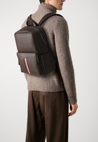 Sac à dos en cuir marron foncé avec une texture lisse, doté d'une poche avant ornée d'un détail rayé rouge et blanc. Bretelles réglables.