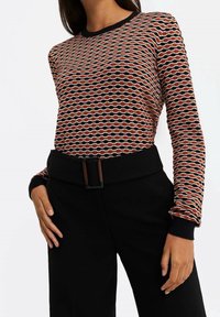 Neulottu pitkähihainen paita, jossa on aaltoileva kuvio oranssin, mustan ja kermaan sävyissä, mustat resorireunukset kaula-aukossa ja hihansuissa. Yhdistetty mustiin housuihin.