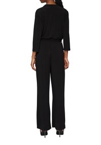 Czarny kombinezon z długimi rękawami, luźny krój, elastyczny pas i szerokie nogawki. Gładka faktura, bez wyraźnych wzorów czy ozdób.