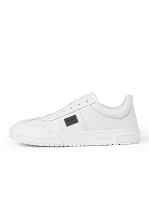 Lage volledig witte sneaker met vetersluiting, gestructureerde zool en zwarte rechthoekige logo patch op het zijpaneel.