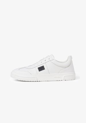 Lage volledig witte sneaker met vetersluiting, gestructureerde zool en zwarte rechthoekige logo patch op het zijpaneel.