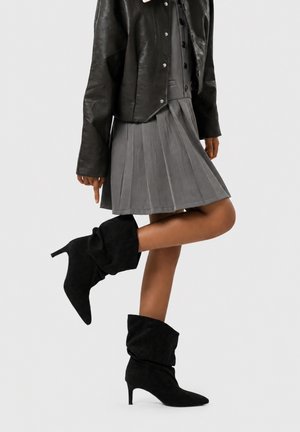 Persona que lleva una chaqueta de cuero negra, falda plisada gris y botas negras de ante con tacón alto, de pie con una pierna doblada.