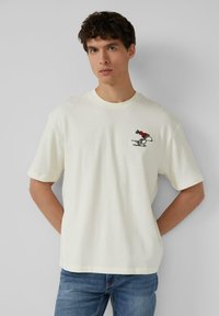 Junger Mann trägt ein weißes T-Shirt mit kleiner Snoopy-Stickerei, eine rote Wintermütze und dazu blaue Jeans. Er steht vor einem hellgrauen Hintergrund.