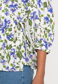Blusa floreale in tessuto leggero con fiori viola e foglie verdi su uno sfondo bianco, con dettagli a fiocco in vita e maniche a sbuffo.
