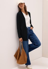 Zwarte cardigan, wit T-shirt, blauwe spijkerbroek, beige sneakers. Draagt een crème pet en houdt een bruine suède schoudertas vast. Casual outfit.