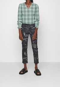 Chemisier à carreaux verts et blancs avec un décolleté en V et des manches bouffantes ; pantalon noir avec texte et graphiques blancs ; sandales à glisser noires.