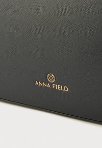 Juodas tekstūruotas paviršius su iškilusiais auksinės spalvos žodžiais „ANNA FIELD“ ir dekoratyviniu apskritu logotipu, sukuriant kontrastuojantį akcentą.