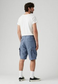 Man wearing white t-shirt, blue cargo shorts, white socks, and black sneakers standing with back turned against plain gray background.