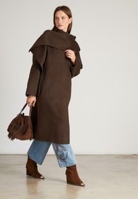 Abrigo de lana marrón con un cuello alto, mangas en capas y un corte relajado. Combinado con jeans de mezclilla azul claros con bordado y botas de tacón marrones.