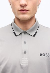 Κοντινό πλάνο ενός ατόμου που φοράει ανοιχτό γκρι πόλο πουκάμισο με γιακά με μαύρο περίγραμμα και το λογότυπο "BOSS" στο στήθος, κουμπωμένο στο λαιμό.