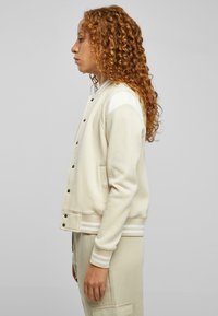 Kremfarget sweatshirt med teksturert overflate, med svarte trykknapper, stripete mansjetter og sidelommer. Krøllete hår synlig.