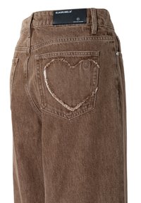 Brune denimbukser har et sydd hjerte-design på baklommen, med en teksturert overflate og merkelapp på midjebåndet.