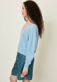 Pull en tricot bleu clair avec un col en V et une coupe oversize ; porté avec un short en denim avec une étiquette sur la poche arrière.