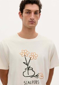 Crème-kleurige katoenen t-shirt met korte mouwen, voorzien van een geborduurd ontwerp van twee bloemen en een vaas in oranje en zwart.