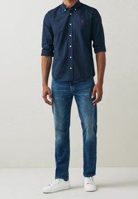 Chemise bleu marine à boutons avec un petit logo rouge, manches longues, associée à un jean bleu clair et des baskets blanches. Coupe décontractée et design simple.