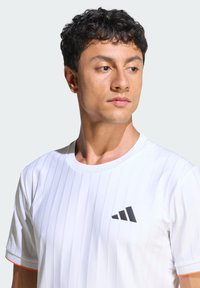 T-shirt de sport blanc à rayures verticales, col rond, avec un logo noir sur le côté gauche de la poitrine. Accents orange sur les manches. Tissu doux.