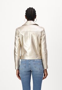 Jachetă din piele metalizată aurie cu guler clasic, design ajustat și accente cu nasturi la talie; combinată cu blugi din denim albastri.