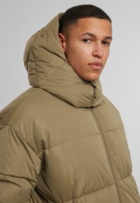 Puffjakke i dempet olivengrønn, med høy krage og quiltet tekstur. Lukkes med glidelås foran og har et minimalistisk design.