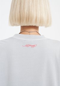 Světlomodré bavlněné tričko s malým dekorativním červeným logem "Ed Hardy" vyšitém na zadní straně poblíž výstřihu. Hladká textura.