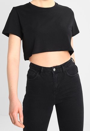 Person trägt ein schwarzes, kurzes Shirt mit kurzen Ärmeln und schwarze High-Waist-Jeans vor einem einfarbigen grauen Hintergrund.