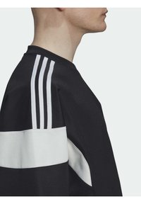 Svart sweatshirt med vita accenter. Har tre vita ränder på axlarna och en kontrastpanel vid midjan. Mjuk textur.