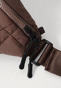 Smeđi kvadratni materijal s crnim zatvaračem i povlačnim trakama; uključuje tamnosmeđu traku s teksturiranim završetkom i metalnim akcentima.
