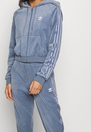 Osoba nosząca niebieski welurowy dres Adidas z kapturem i kurtką na suwak oraz dopasowane spodnie z białymi paskami na rękawach i nogawkach.