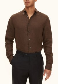 Camicia di lino marrone con colletto button-down, maniche lunghe e texture del tessuto. Abbinata a pantaloni scuri su misura. Presenta bottoni argentati.