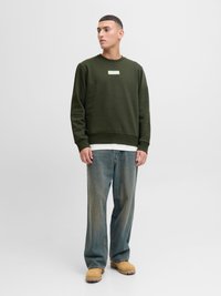 Mand stående iført en olivengrøn sweatshirt, hvid undertrøje, løse blå jeans og brune arbejdsstøvler, kigger til siden mod en ensfarvet baggrund.
