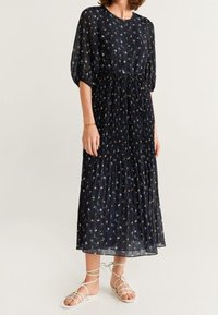 Robe bleu marine à motif floral avec des manches bouffantes courtes, taille froncée et jupe fluide à plusieurs niveaux. Portée avec des sandales à lanières.
