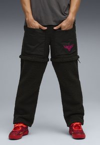 Pantaloni in pile nero con grandi tasche nere, dettaglio logo rosa e superficie testurizzata e pelosa. Abbinati a scarpe rosso acceso.