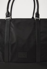 Black tote bag made of nylon with leather straps, featuring a rectangular logo patch. Smooth texture with metal hardware accents.
