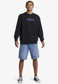 Schwarzer Hoodie mit blauem Grafikdesign, kombiniert mit hellblauen Jeansshorts. Das Modell trägt schwarze und weiße Sneakers. Lässiges Outfit.