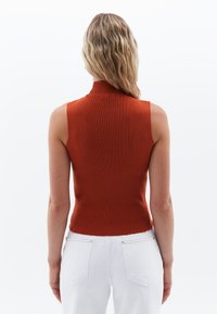 Top senza maniche color ruggine con collo alto, texture a coste e design aderente, abbinato a jeans in denim bianco.