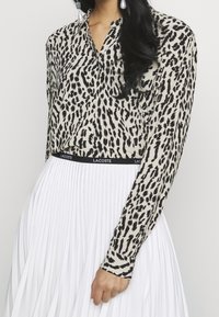 Mujer con una blusa de leopardo de manga larga metida dentro de una falda blanca plisada con una cinturilla negra de Lacoste, sosteniendo el borde de la falda.