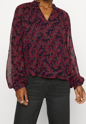 Person trägt eine marineblaue Bluse mit rotem Blumenmuster, gerafftem Kragen und transparenten langen Ärmeln kombiniert mit dunkelgrauer Hose.