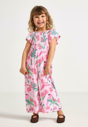 Ragazza giovane sorridente con capelli ricci, indossa un vestito rosa con motivi floreali e di uccelli e scarpe intrecciate marroni, in piedi su uno sfondo bianco semplice.