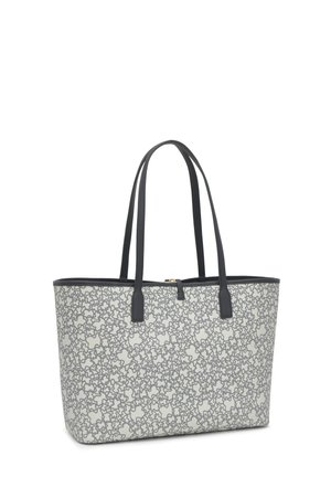 Persona sosteniendo una gran bolsa tote blanca con patrones de flores grises y asas negras, vistiendo ropa de mezclilla azul oscuro y botas negras con tacón.