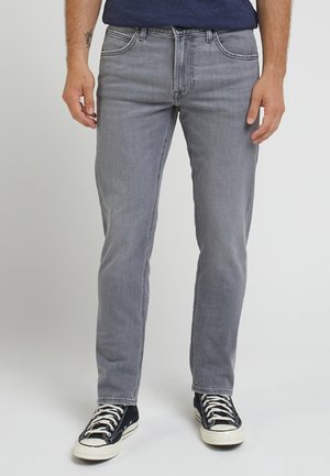 Hombre con vaqueros de corte ajustado gris y zapatillas negras bajas de pie frente a un fondo liso.